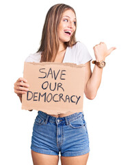 Young beautiful blonde woman holding save our democracy protest banner pointing thumb up to the...