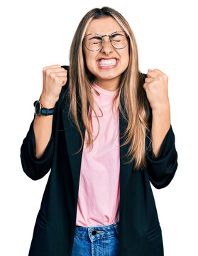 Hispanic young woman wearing business jacket and glasses excited for success with arms raised and eyes closed celebrating victory smiling. winner concept.