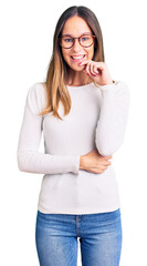 Beautiful brunette young woman wearing casual white sweater and glasses looking confident at the camera smiling with crossed arms and hand raised on chin. thinking positive.
