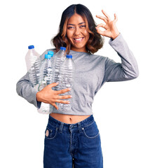Young beautiful mixed race woman holding recycling plastic bottles doing ok sign with fingers, smiling friendly gesturing excellent symbol