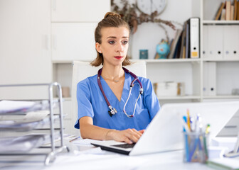 Cheerful woman doctor sitting at workplace with computer in her office