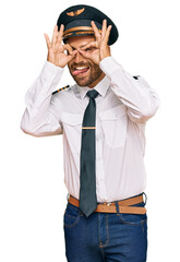 Handsome man with beard wearing airplane pilot uniform doing ok gesture like binoculars sticking tongue out, eyes looking through fingers. crazy expression.