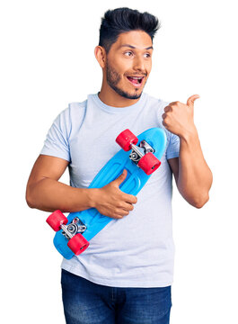 Handsome latin american young man holding skate pointing thumb up to the side smiling happy with open mouth