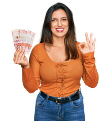 Beautiful hispanic woman holding 10 colombian pesos banknotes doing ok sign with fingers, smiling...