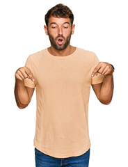 Handsome young man with beard wearing casual tshirt pointing down with fingers showing advertisement, surprised face and open mouth