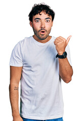 Hispanic young man with beard wearing casual white t shirt surprised pointing with hand finger to the side, open mouth amazed expression.