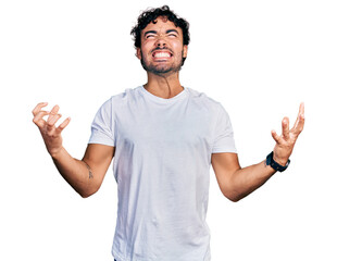 Hispanic young man with beard wearing casual white t shirt crazy and mad shouting and yelling with aggressive expression and arms raised. frustration concept.