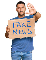 Handsome man with tattoos holding fake news banner with open hand doing stop sign with serious and...