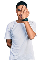Fototapeta premium Young hispanic man wearing casual white t shirt covering one eye with hand, confident smile on face and surprise emotion.