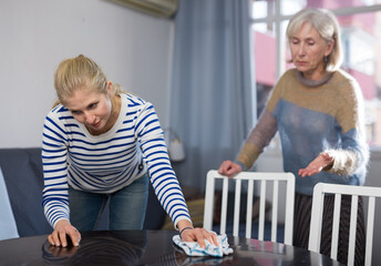 Mature woman scolds her adult daughter, wiping the table in the room with a rag