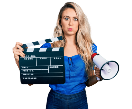 Young blonde woman holding video film clapboard and megaphone puffing cheeks with funny face. mouth inflated with air, catching air.