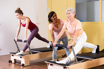 Fototapeta na wymiar Two health conscious women of different ages perform a Pilates exercise using a reformer bed, where a female instructor ..helps them do it correctly