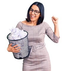 Young beautiful latin woman holding paper bin full of crumpled papers screaming proud, celebrating victory and success very excited with raised arms