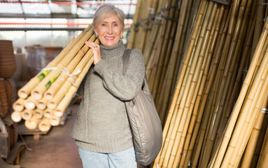 Smiling elderly woman carrying on shoulder bunch of bamboo poles bought at horticultural market for landscaping garden..