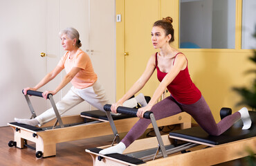 Fototapeta na wymiar Elderly woman and young girl practicing exercises on Pilates reformer. Concept of pilates as physical therapy alternative for injury rehab