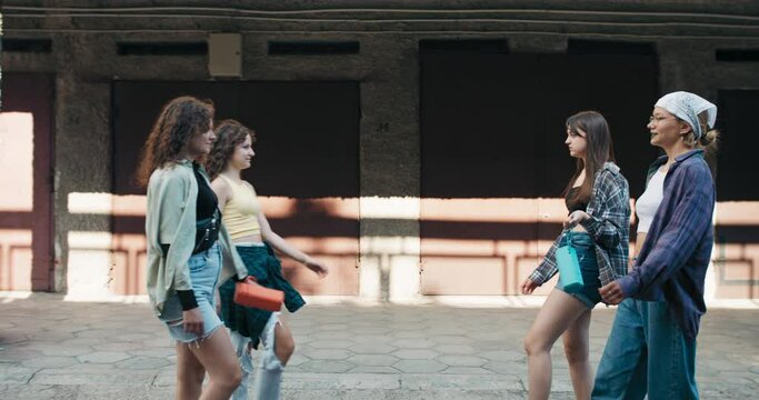 A group of girls meet in neighborhood garages in the city to do a dueling dance. The teens hold portable music speakers in their hands.