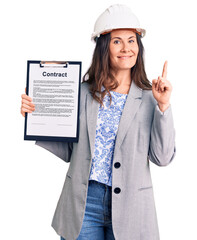Young beautiful brunette woman wearing architect hardhat holding contract surprised with an idea or question pointing finger with happy face, number one
