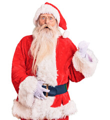 Old senior man with grey hair and long beard wearing traditional santa claus costume surprised pointing with hand finger to the side, open mouth amazed expression.