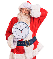 Old senior man with grey hair and long beard wearing santa claus costume holding clock stressed and frustrated with hand on head, surprised and angry face