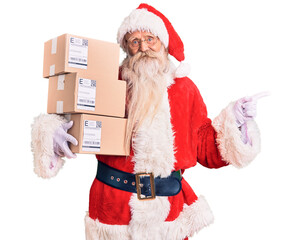 Old senior man with grey hair and long beard wearing santa claus costume holding boxes smiling happy pointing with hand and finger to the side - Powered by Adobe