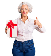 Senior grey-haired woman holding gift smiling happy and positive, thumb up doing excellent and approval sign