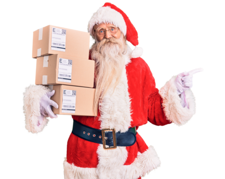 Old senior man with grey hair and long beard wearing santa claus costume holding boxes smiling happy pointing with hand and finger to the side