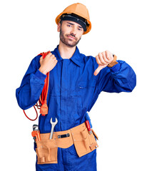 Young hispanic man wearing elecrician uniform holding cable with angry face, negative sign showing dislike with thumbs down, rejection concept