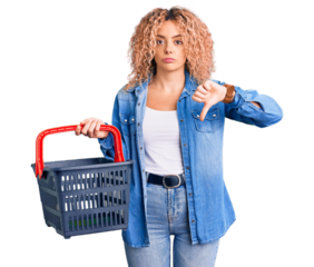 Young blonde woman with curly hair holding supermarket shopping basket with angry face, negative sign showing dislike with thumbs down, rejection concept