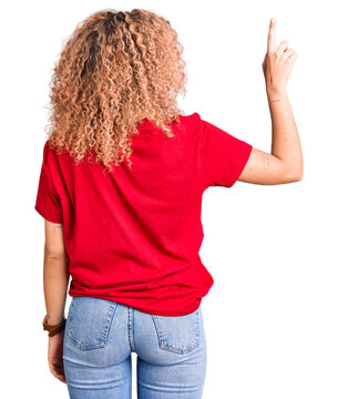 Young blonde woman with curly hair wearing casual red tshirt posing backwards pointing ahead with finger hand