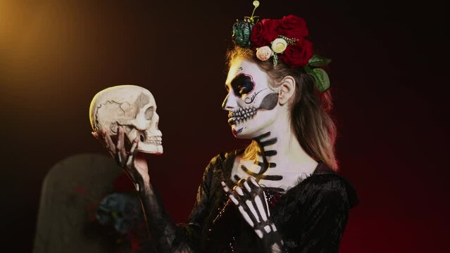 Horror santa muerte looking at skull in studio, wearing creepy halloween costume with headband. Acting like holy glamorous entity on dios de los muertos, having black and white make up. Handheld shot.