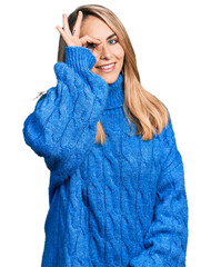 Young blonde woman wearing casual sweater doing ok gesture with hand smiling, eye looking through fingers with happy face.