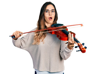 Beautiful hispanic woman playing violin afraid and shocked with surprise and amazed expression, fear and excited face.