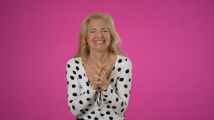 Excited surprised overjoyed happy blonde woman 50s wears polka dot shirt doing winner gesture celebrate clenching fists say yes isolated on solid pink background studio portrait