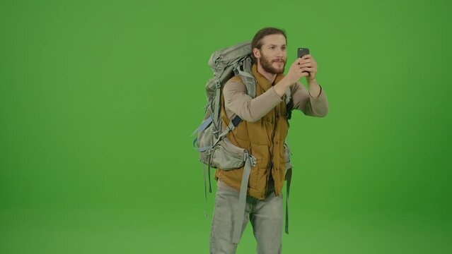 Young Inspired Bearded Man with Big Travel Backpack Looking Around the Beautiful Views in Front of Him, Filming It Wondered on Smartphone,Making Selfie, Photos and Smiling on Green Screen,Chroma Key,