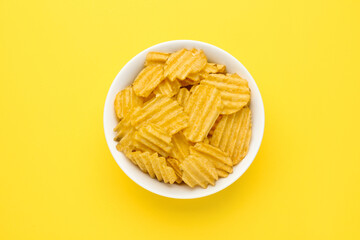 Bowl of tasty ridged potato chips on yellow background, top view