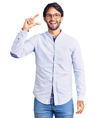 Handsome hispanic man wearing business shirt and glasses smiling and confident gesturing with hand doing small size sign with fingers looking and the camera. measure concept.