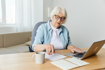 an elderly woman sitting at a laptop and working, from home in her cozy apartment, with a cup and notebooks on the table