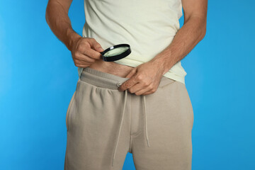 Man examining intimate area with magnifying glass on light blue background, closeup. Genital herpes