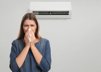 Woman suffering from cold in room with air conditioner on white wall, space for text