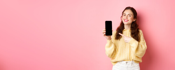 Portrait of attractive young woman demonstrates promo on phone, pointing finger at smartphone and...