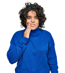 Young hispanic woman with curly hair wearing turtleneck sweater touching mouth with hand with...