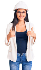 Young beautiful latin girl wearing architect hardhat and glasses success sign doing positive gesture with hand, thumbs up smiling and happy. cheerful expression and winner gesture.