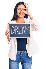 Young beautiful latin girl holding blackboard with dream word smiling happy doing ok sign with hand on eye looking through fingers