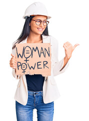 Young beautiful latin girl wearing architect hardhat holding woman power banner pointing thumb up to the side smiling happy with open mouth