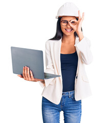 Young beautiful latin girl wearing architect hardhat holding laptop smiling happy doing ok sign with hand on eye looking through fingers