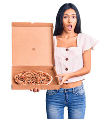 Young beautiful latin girl holding delivery pizza box scared and amazed with open mouth for...