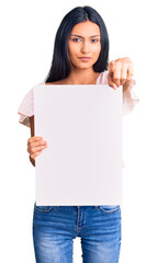 Young beautiful latin girl holding blank empty banner pointing with finger to the camera and to you, confident gesture looking serious