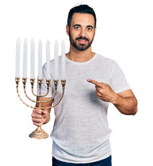 Young hispanic man with beard holding menorah hanukkah jewish candle smiling happy pointing with...