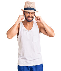Young hispanic man wearing casual summer hat covering ears with fingers with annoyed expression for the noise of loud music. deaf concept.