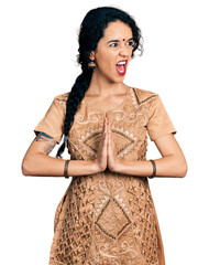 Young indian woman doing anjali mudra angry and mad screaming frustrated and furious, shouting with...